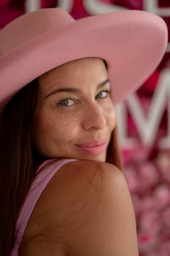 A Starseed Summit guest in a pink hat wearing Fazit Beauty face sparkles against a rose wall