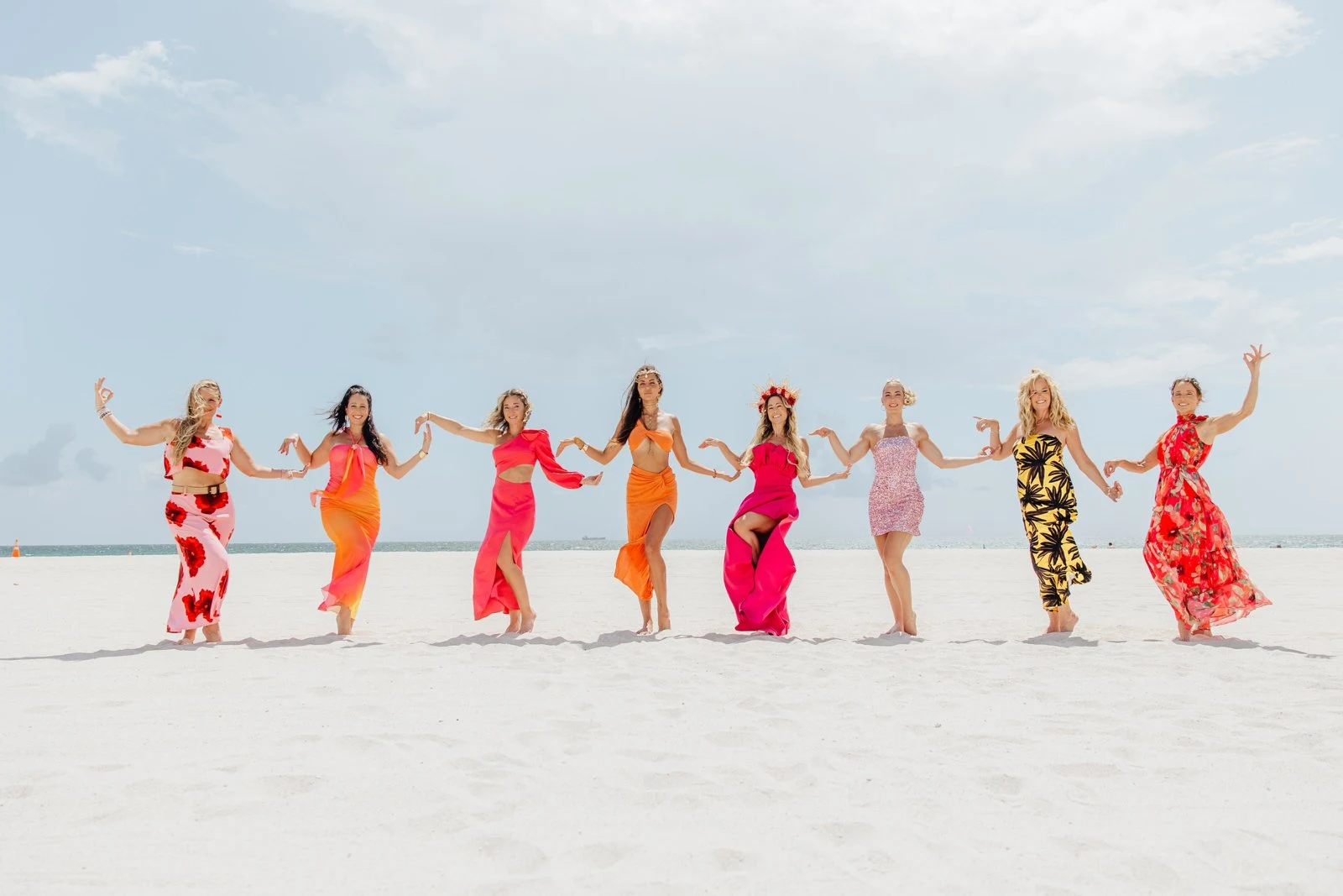 Eight Starseed Summit guests in coral and hot-pink gowns, hand in hand along the sand at South Beach